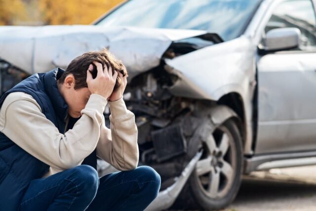 A distressed man stands near a heavily damaged car, holding his head in frustration as he realizes the car is beyond repair.