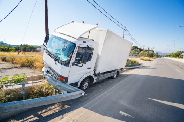 A crashed truck that hit a damaged road barrier, with a broken windshield, on a sunny day.