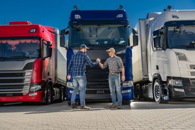 Two Caucasian semi-truck drivers shaking hands in front of a group of European trucks.